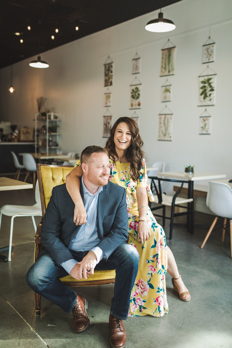 A smiling couple seated together, with the woman standing behind the man in a floral dress, captured by Luke and Ashley Photography, in a modern café with hanging botanical artworks.