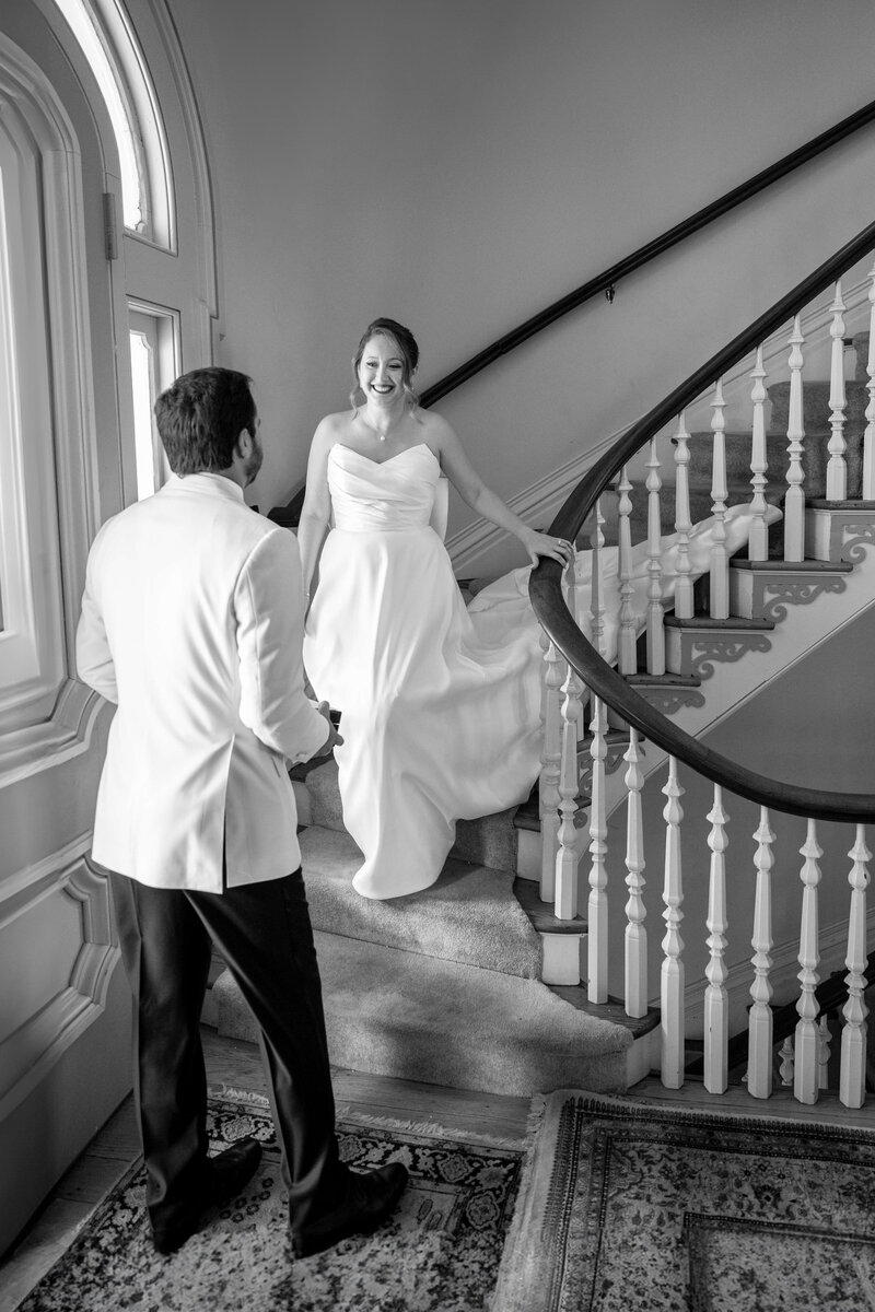 A bride in a white gown descends a curved staircase, smiling at a groom in a white jacket. Both appear excited, standing in a well-lit, elegant interior with large windows.