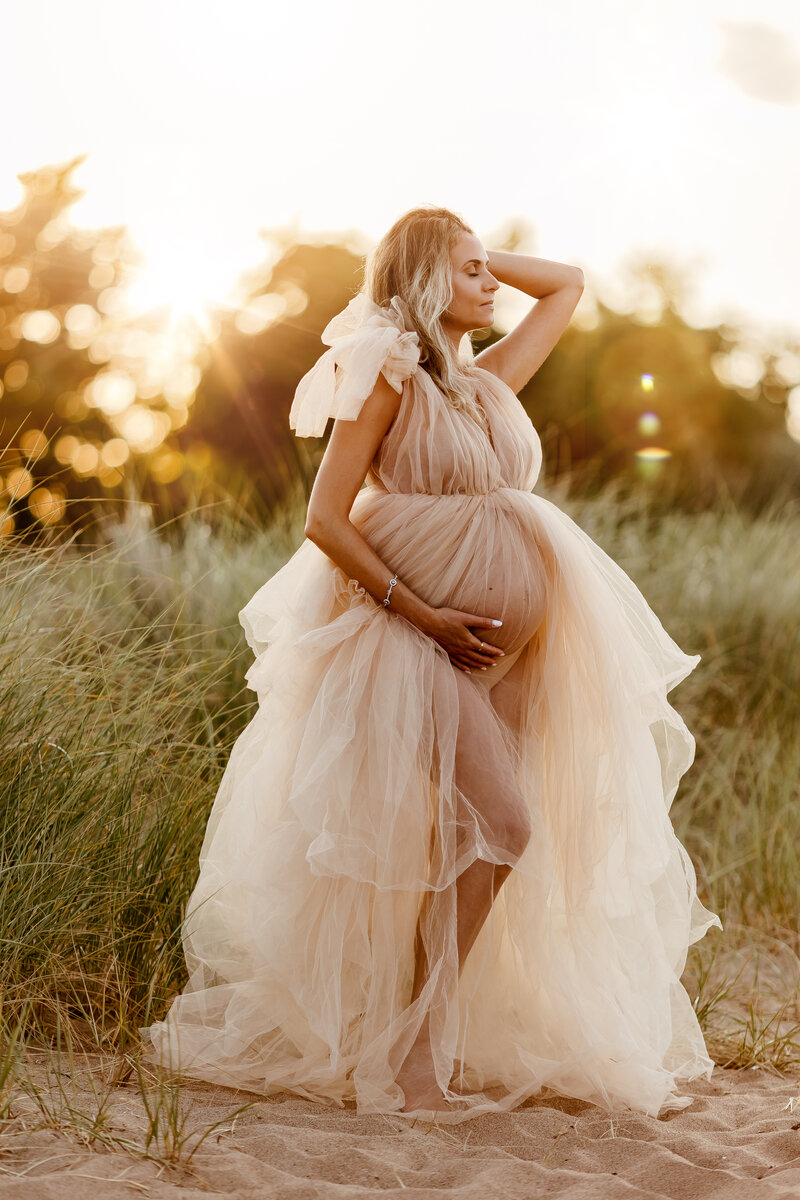 pregnant woman standing on the beach with left arm in her hair, right hand hugging her belly, with her eyes closed and the sunset light around her 