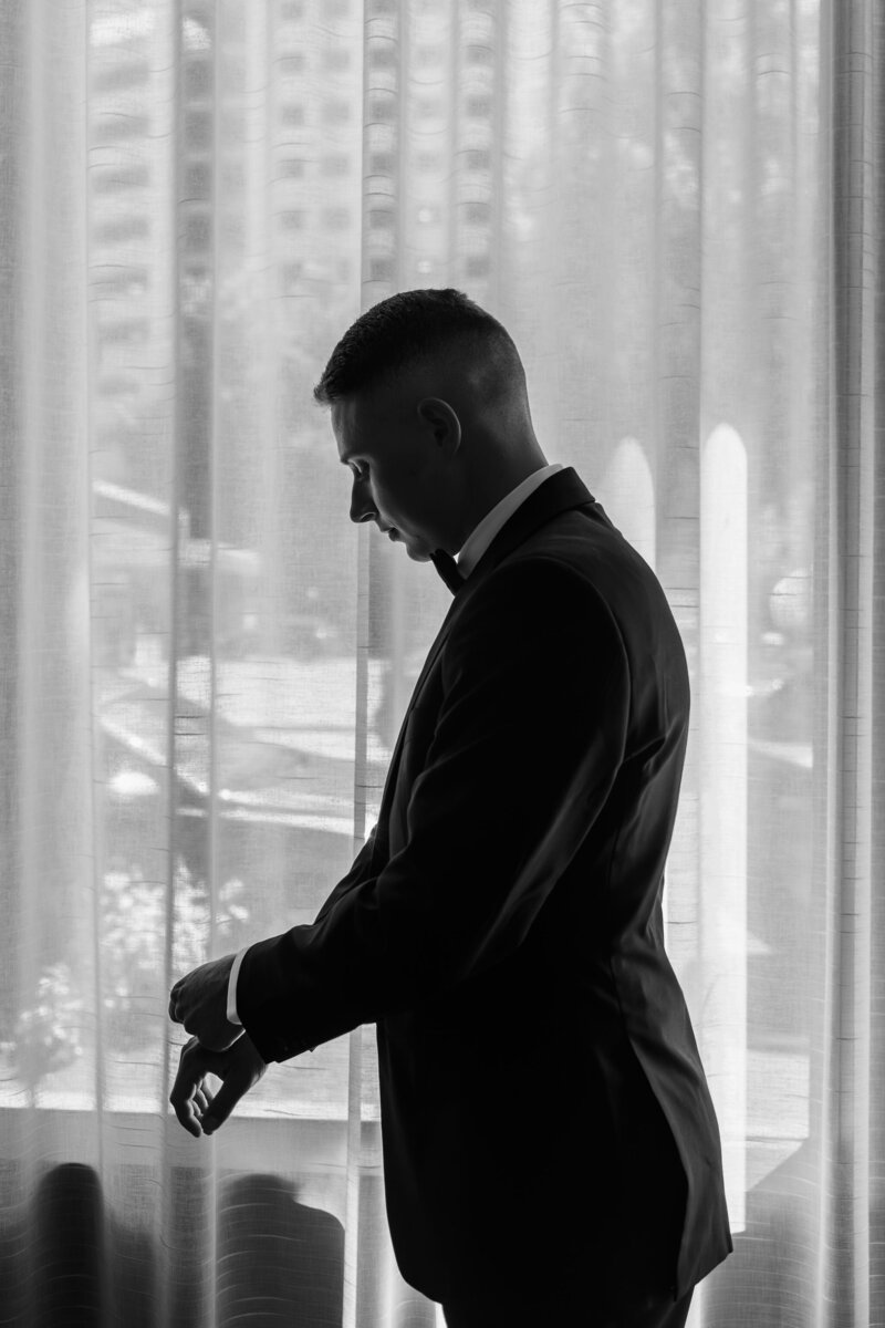 Black and white silhouette of a person in a suit checking their watch by a window with sheer curtains and the downtown Portland cityscape in the background, capturing a timeless pre-wedding moment.