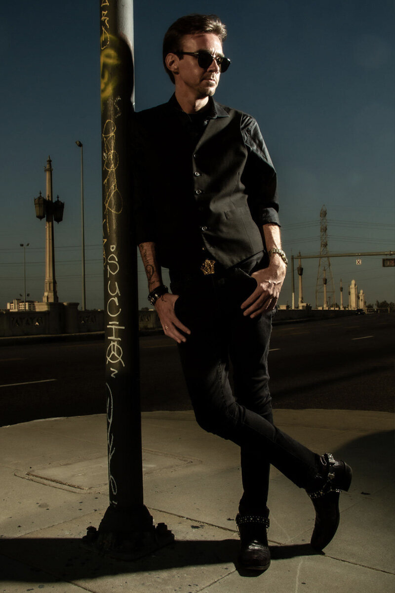 Professional Branding Photo Chandler Morrison standing against street lamp wearing sunglasses thumbs in pockets on leg crossed tip of boot on pavement