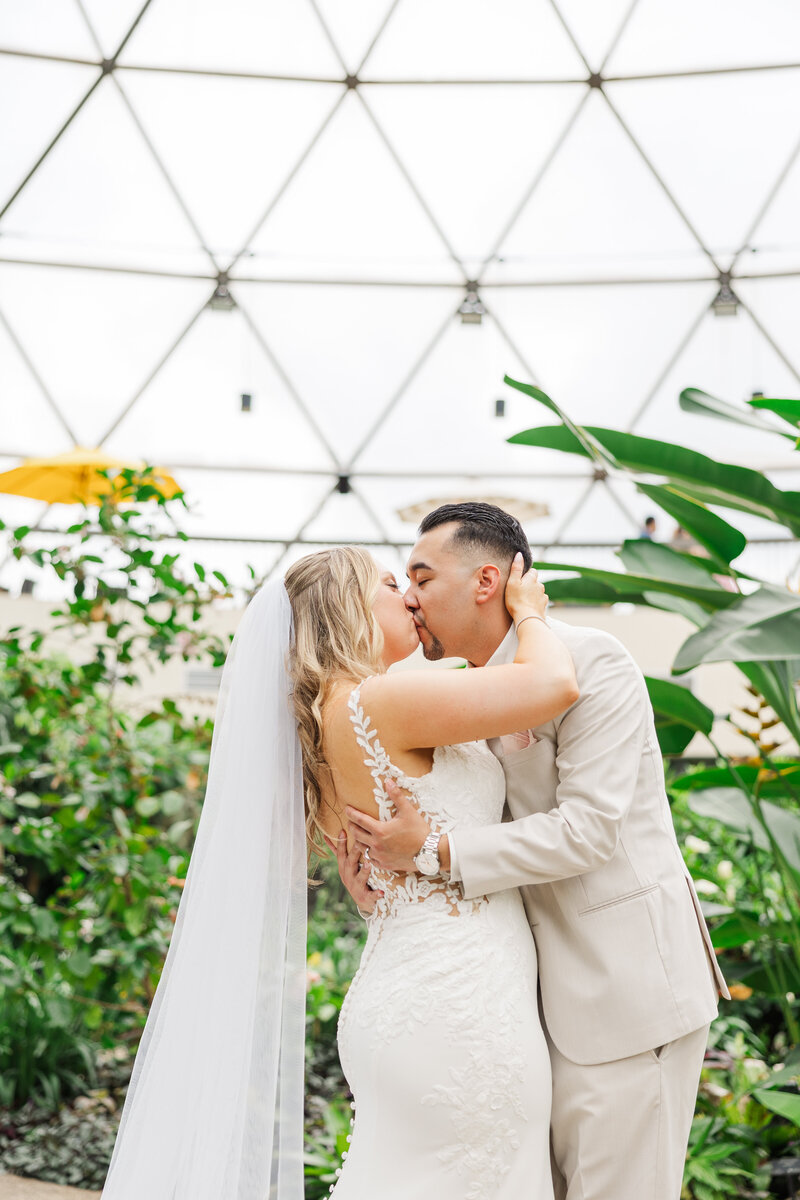 A couple kisses under the dome of the Greater Des Moines Botanical Garden in this destination wedding pic by Claire Katan.