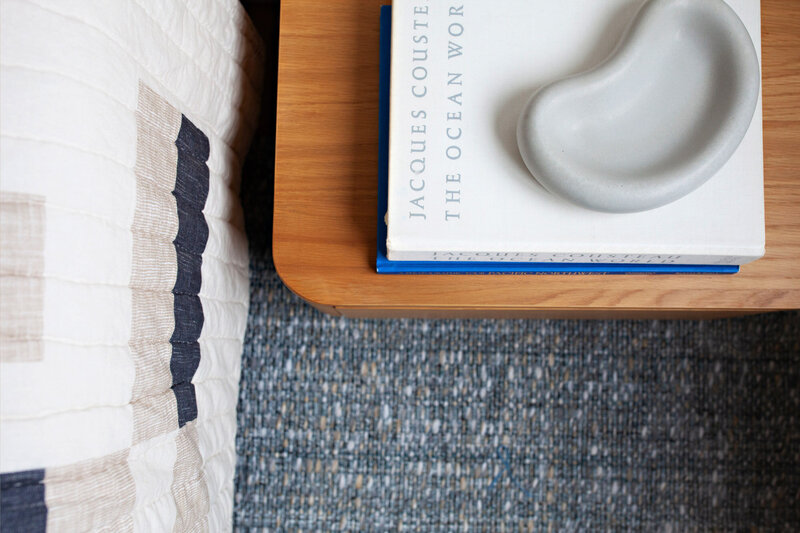 Close-up of a cozy corner in an Oregon beach house rental featuring a striped quilt, a textured blue rug, and a wooden nightstand with a coastal-themed book and decorative tray.