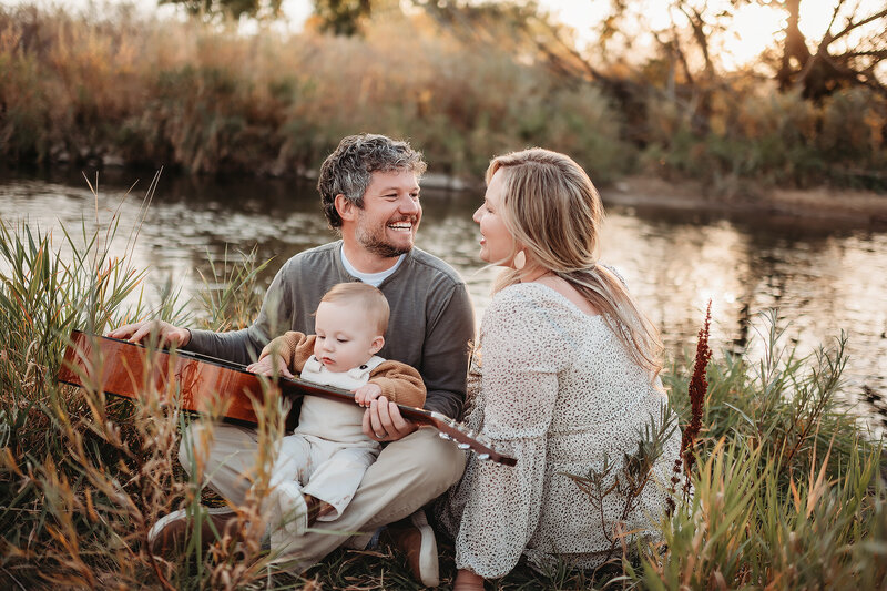 dad holding a guitar in his lap while baby boy strums it and mom looks at dad adoringly beside a river