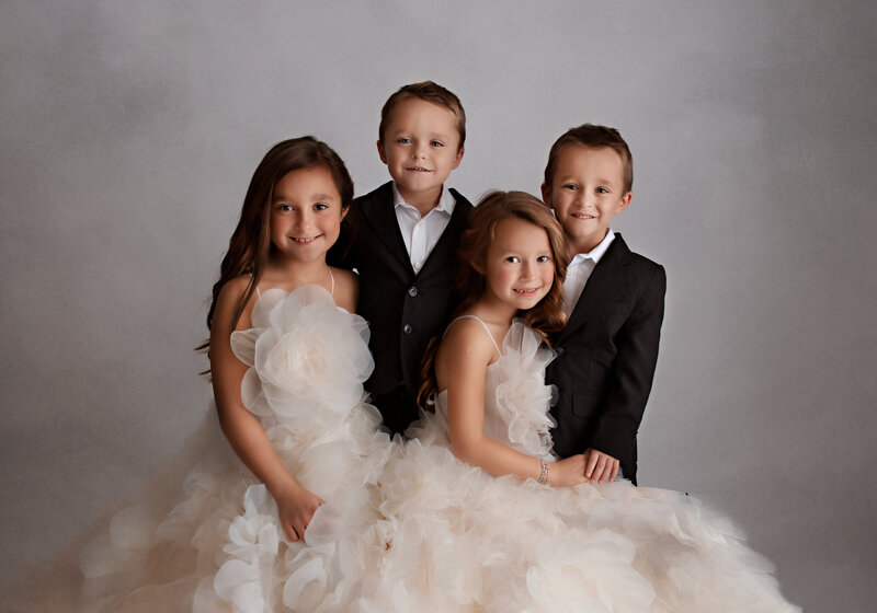 Photo of four children, 2 girls and 2 twin boys dressed in formal attire.  Girls are wearing white flower dresses and the boys are wearing black suits with white shirts