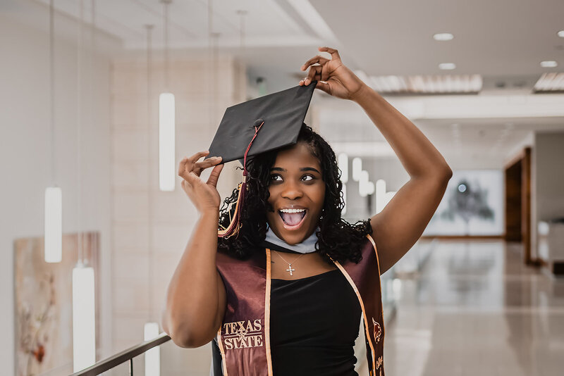 College graduate in black pantsuit at Texas State Campus holding cap on head and smiling.