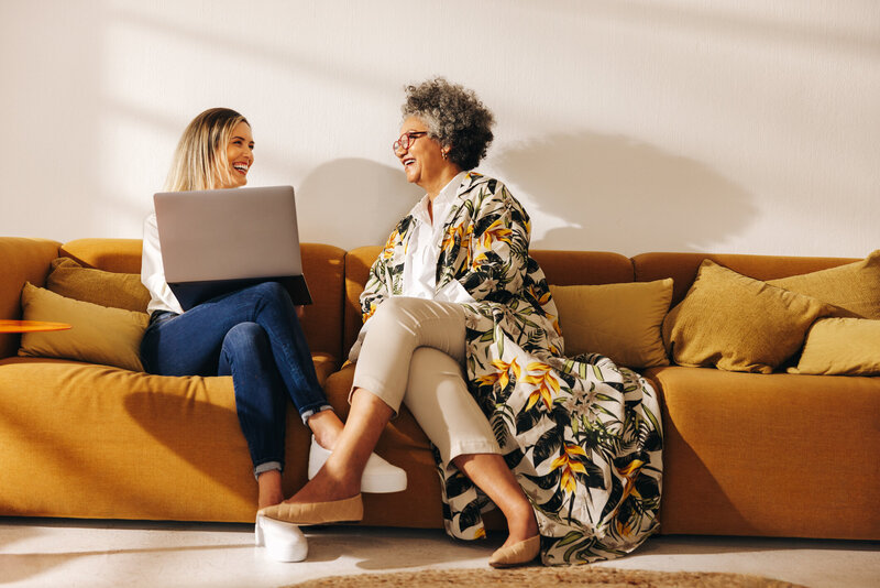 Two women colleagues share a laugh on a couch. The white woman is balancing a laptop. The older Black woman wears glasses and a long floral cardigan