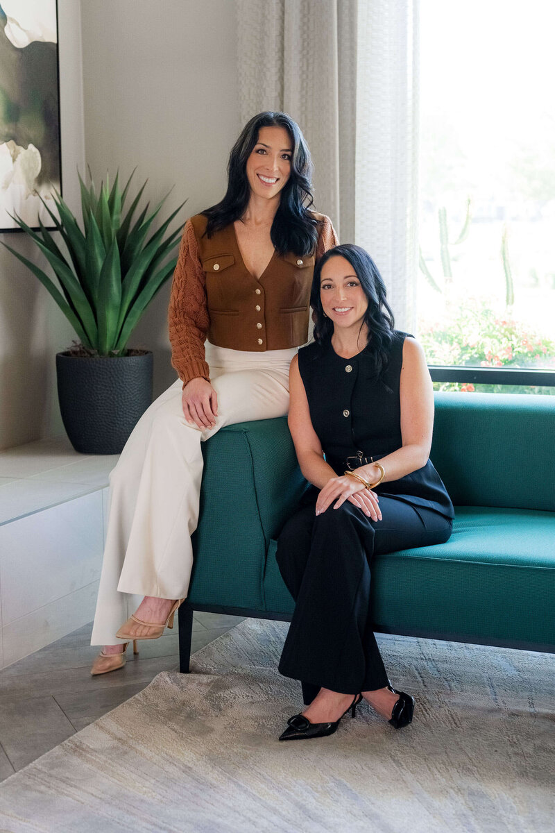 Two women on a couch, engaged in a conversation about sales techniques, with a plant nearby.
