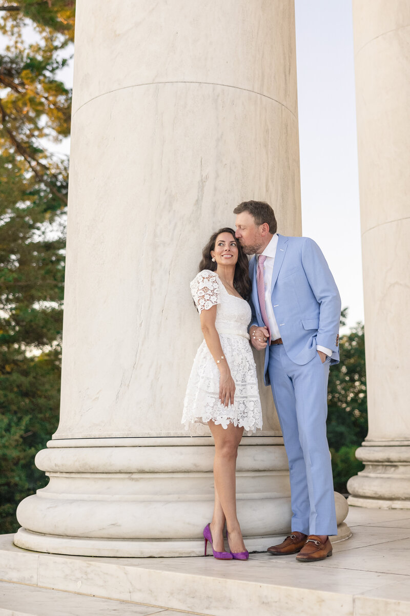 A couple stands in front of a large marble column, embracing and smiling. The woman is wearing a short white lace dress and purple shoes, while the man wears a light blue suit and pink tie. Trees and a clear sky are visible in the background.