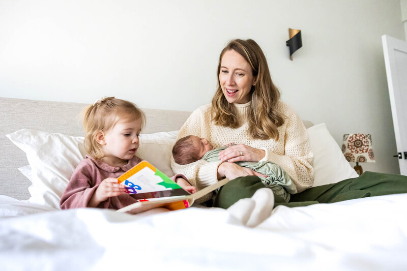 Boston mom sitting on bed with newborn in her arms and reading to toddler daughter