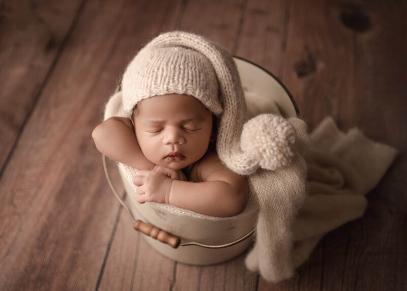 Best Newborn Photographer Lake Elsinore | baby boy resting chin on hands posed in cream bucket with matching cream knit pom pom sleepy hat