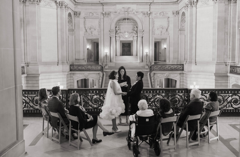 LGBTQ+ couple exchanging vows in an intimate city hall wedding ceremony surrounded by close family, with grand architectural details in the background.