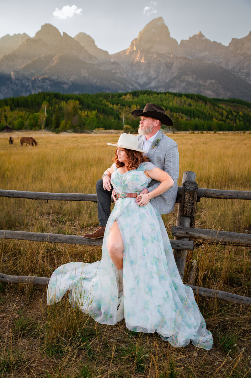 Jackson Hole photographers capture intimate Grand Teton wedding with bride and groom leaning against fence in wedding attire