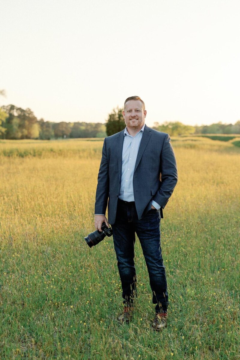 A man holding a camera stands in a field with tall grass during sunset, capturing luxury wedding photography.