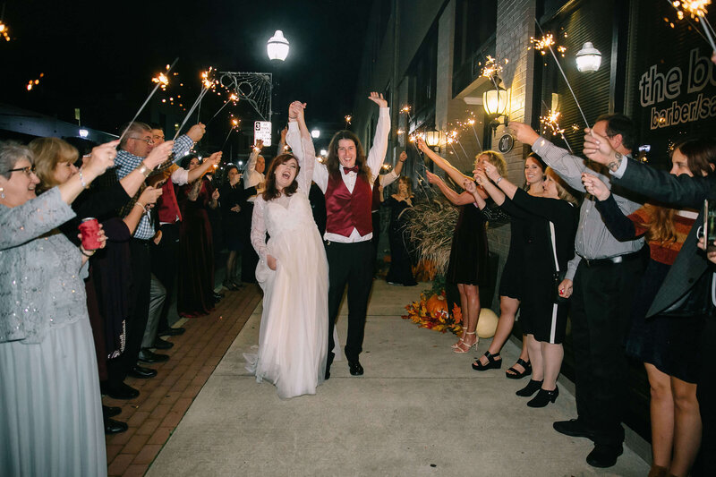A bride and groom celebrate as they walk hand-in-hand through a cheering crowd holding sparklers outside at night. The couple smiles joyfully, surrounded by friends and family lined along the sidewalk.