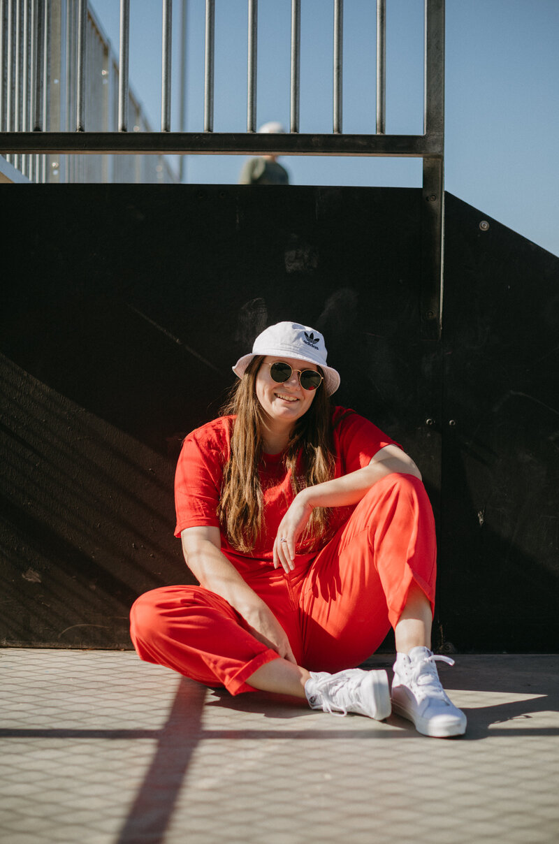 Girl sitting on the ground in a red outfit smiling at a skatepark.