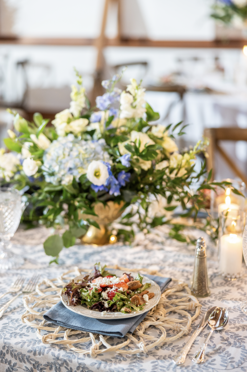 Spring wedding at WinMock at Kinderton, designed by Charming Carolina — table styled with dusty blue patterned linen, a low floral centerpiece, preset salad, wicker placemat, and a dusty blue napkin.