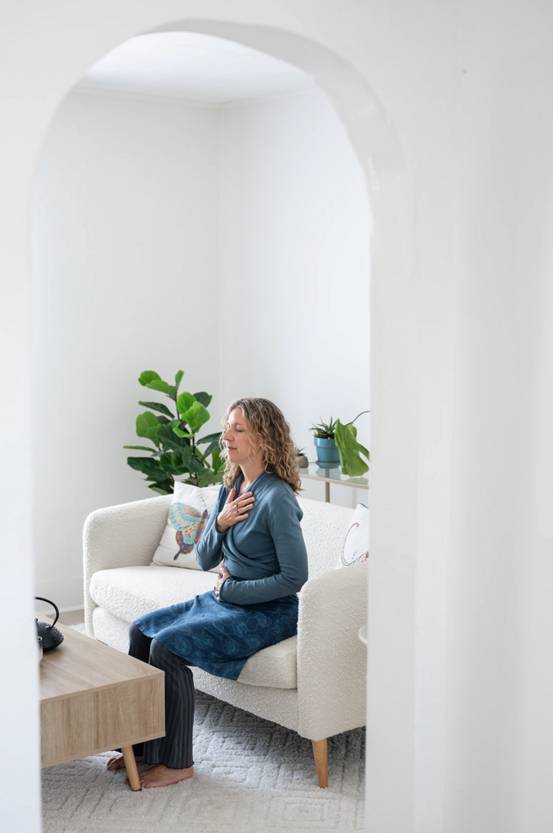 Somatic Experiencing therapist Heidi Schauster sits with her hands on her chest and abdomen. She's surrounded by plants, conveying calmness and introspection.