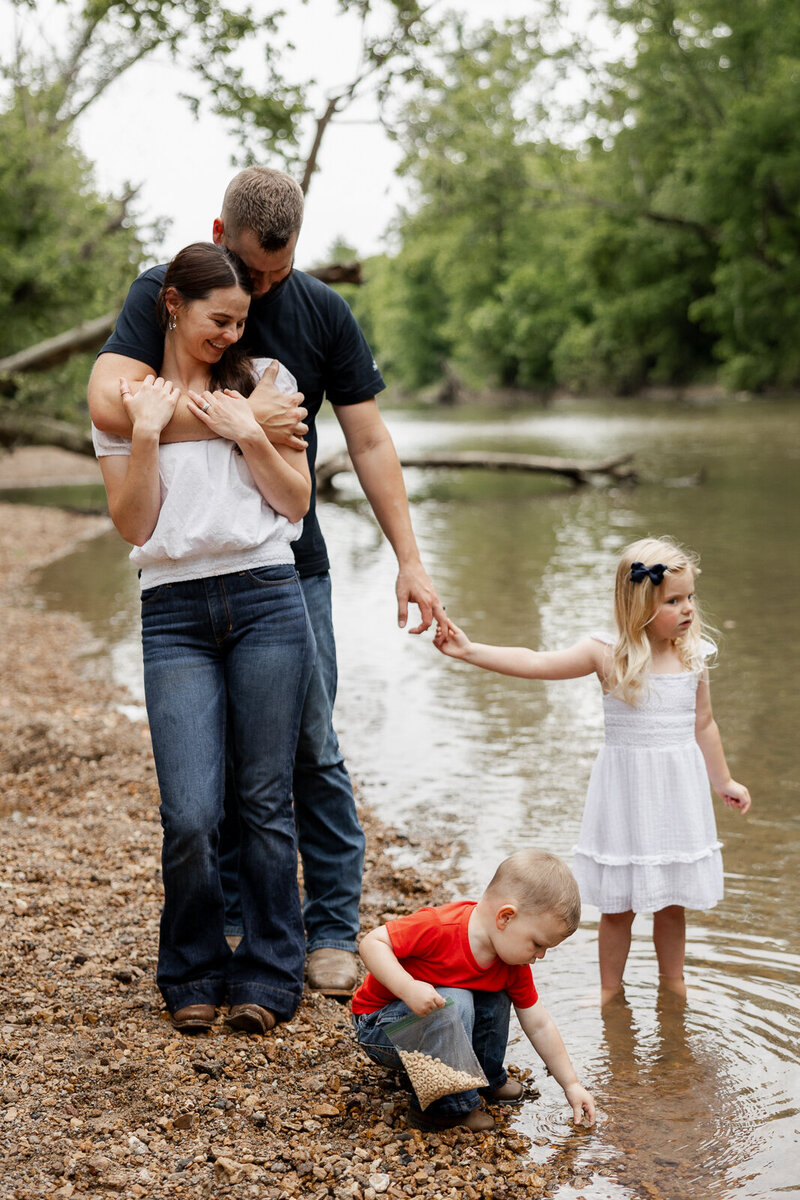 Family portrait at the creek dad hugging mom, son eating cheerios out of a bag reaching into the water and daughter holding dad's hands. 
