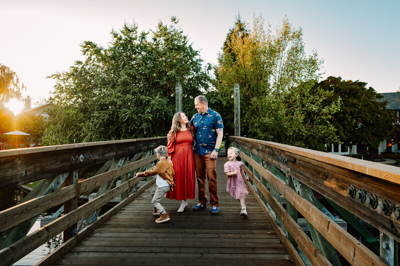 A family of four is captured in a lively lifestyle portrait on a wooden bridge, the sun going down behind them. The parents, standing in the center, share a loving gaze while holding hands. The mother is dressed in a flowing red dress, and the father wears a blue shirt with brown pants. Their two young children, a boy and a girl, are playfully running around them, smiling and enjoying the moment. The background features lush green trees, with sunlight filtering through, creating a serene and joyful atmosphere.