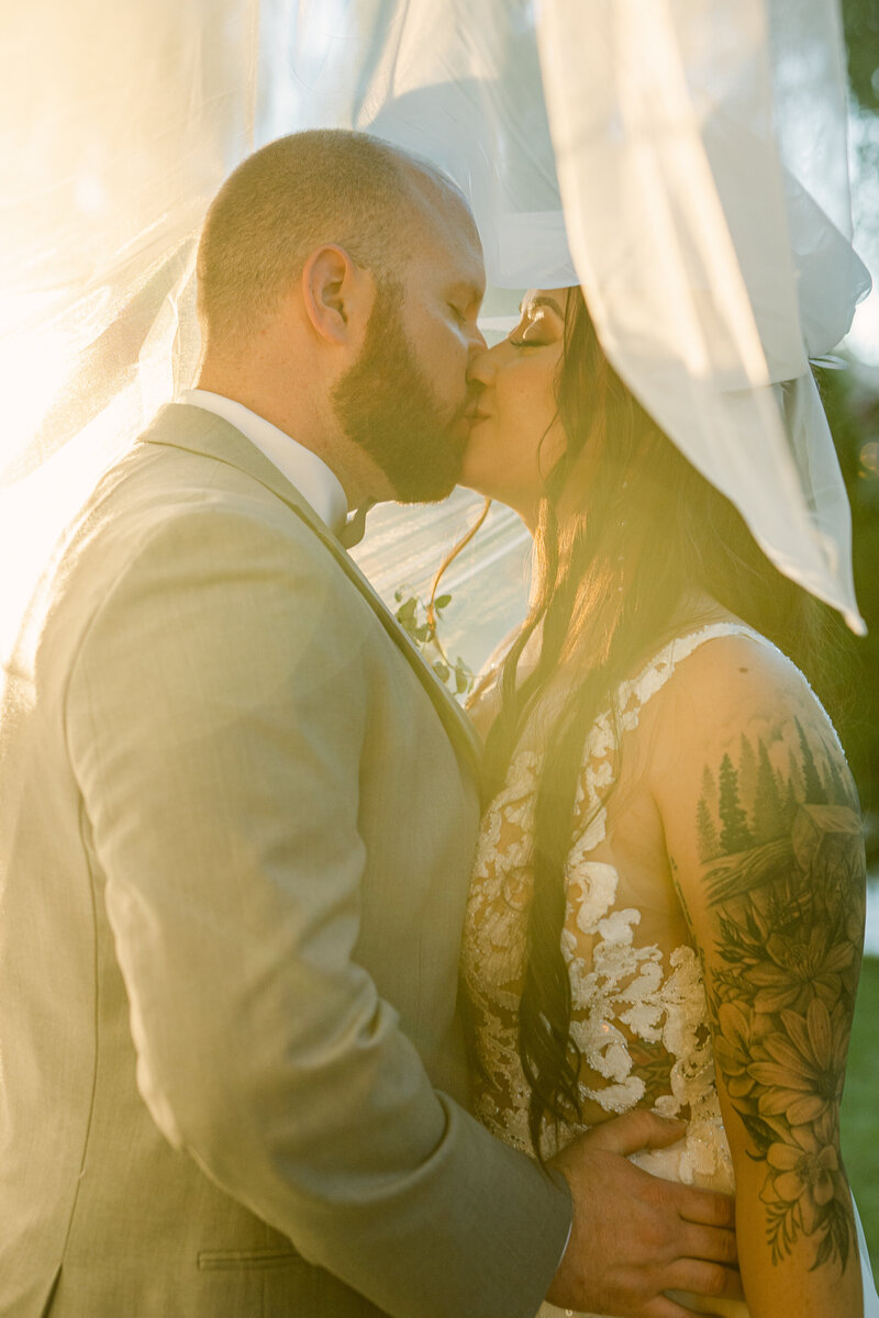 A tattooed bride and groom kissing under a veil at sunset.