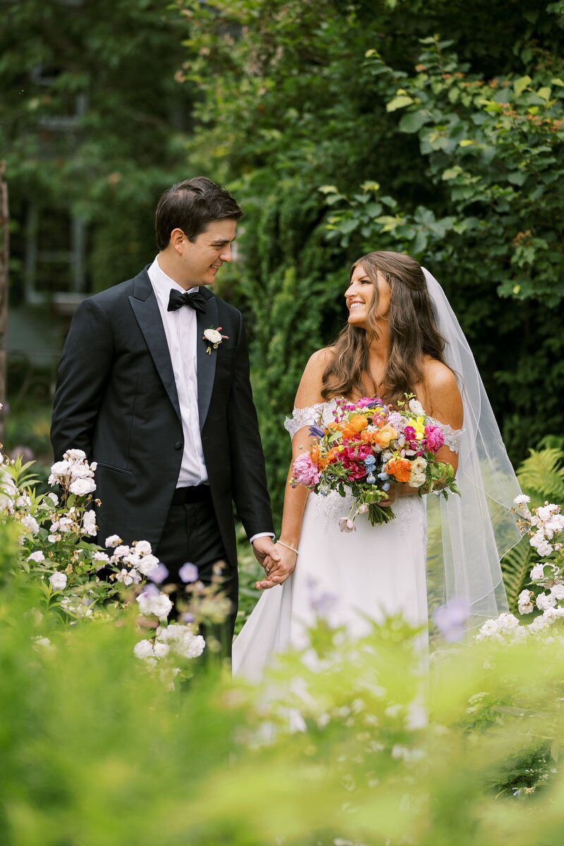 Bride and groom walking hand-in-hand through a lush garden, smiling at each other, with the bride holding a colorful bouquet.
