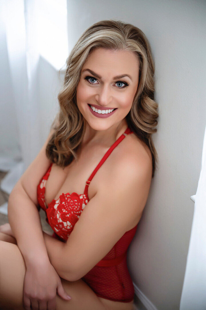 A woman with long hair, wearing a red lace outfit, is seated by a sunlit window. She is smiling warmly, creating a cheerful and confident atmosphere.