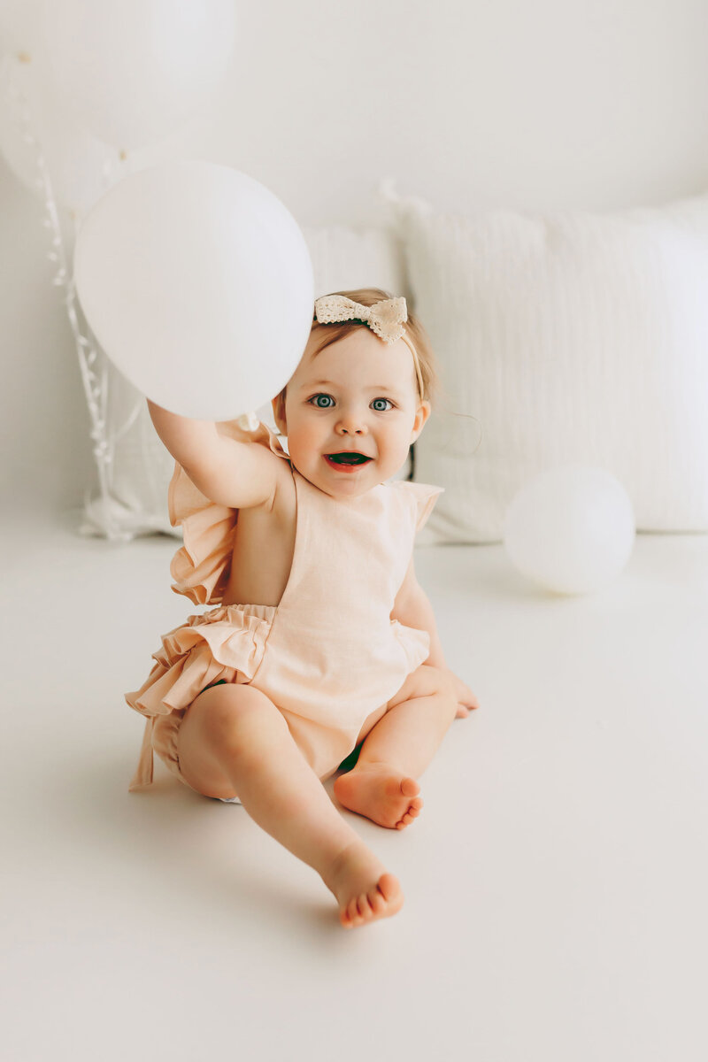 A one year old girl sits playing with a balloon during her first birthday session inside a Vancouver Washington natural light photo studio