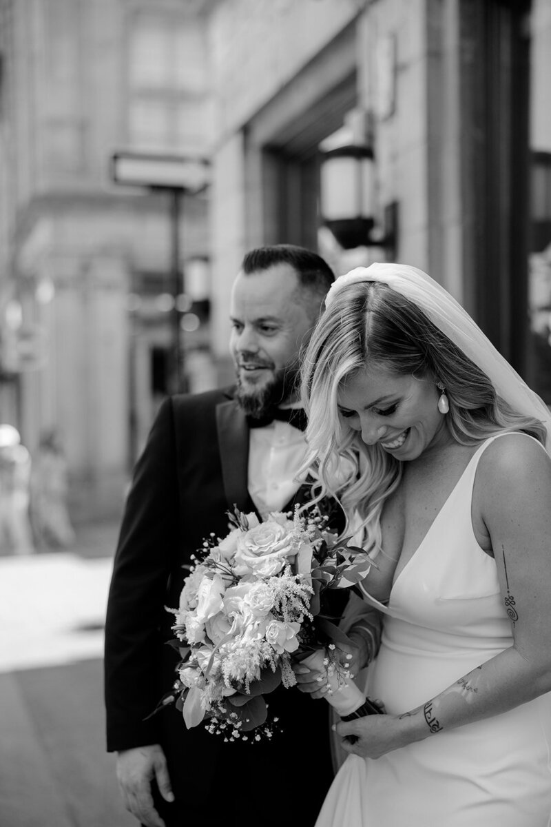 A black and white photograph of a smiling couple on their wedding day in an urban setting.