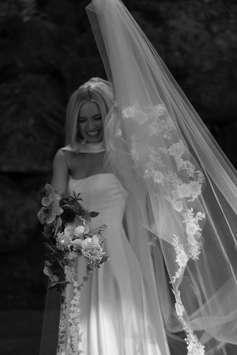 Black and white portrait of smiling bride in flowing veil holding bouquet with trailing florals