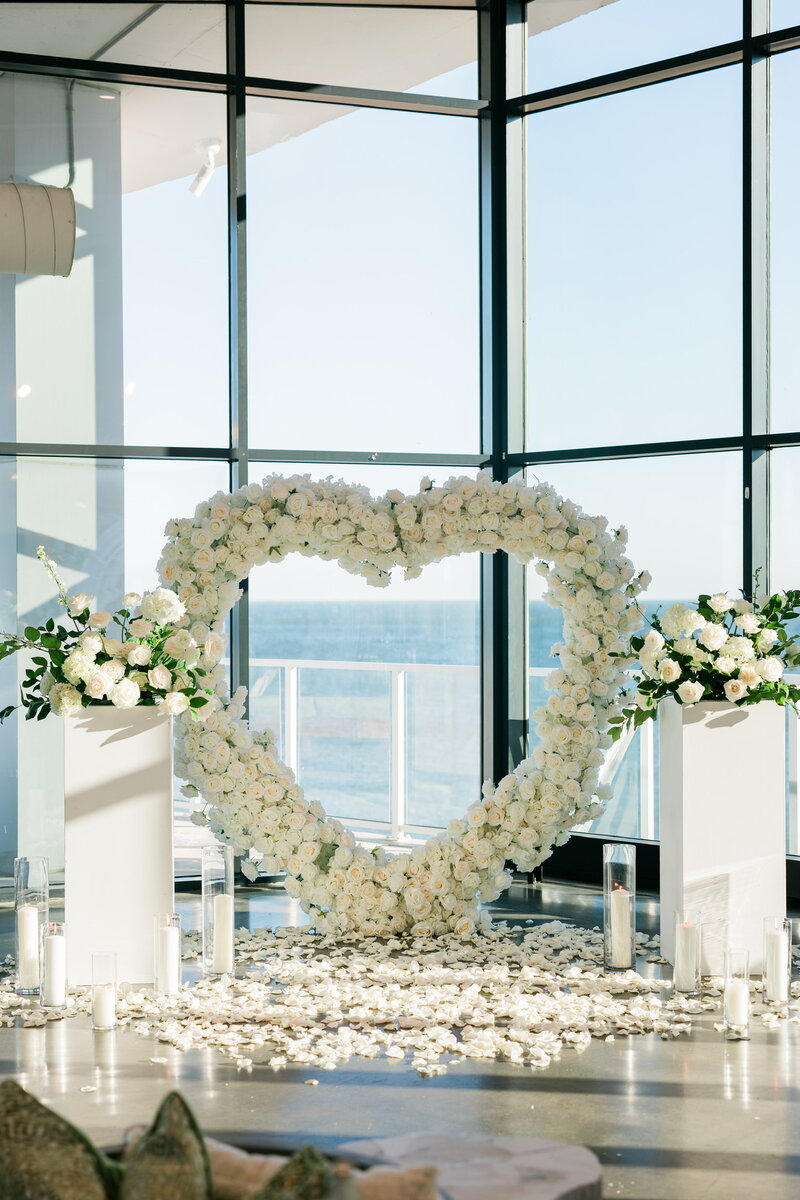 Indoor wedding ceremony backdrop featuring a large white floral heart arch with petal aisle and ocean views, styled by Yours Truly, a wedding planner and designer in New Jersey.