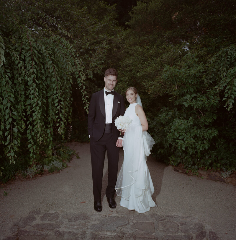 A romantic wedding portrait of the bride and groom taken at Burnley Gardens in Richmond before their Cutler and Co wedding reception. The couple stands side by side on a stone pathway surrounded by lush greenery, creating a serene, natural backdrop. The groom is dressed in a classic black tuxedo with a bow tie, exuding elegance, while the bride looks radiant in a sleek, white halter-neck gown paired with a long, flowing veil. She holds a bouquet of white flowers, adding a delicate touch to her modern, minimalist look. Both are smiling softly, looking content and relaxed as they pose together in the tranquil garden setting, capturing the timeless beauty and joy of their special day.