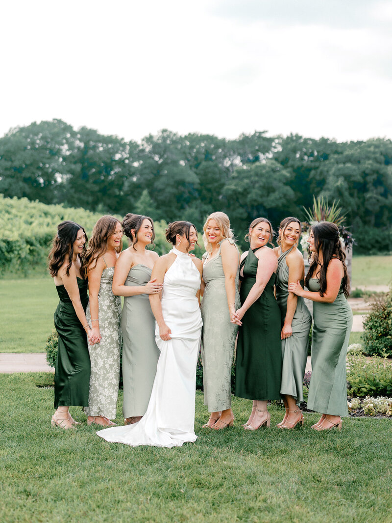 A bride and her bridesmaids in mismatched green dresses standing outside and smiling at each other.