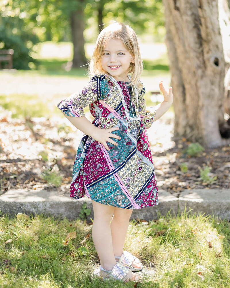 Young girl with blonde hair standing in the grass at a park, wearing a patterned half sleeve dress, standing to the side, and smiling at the camera with a hand on her hip.