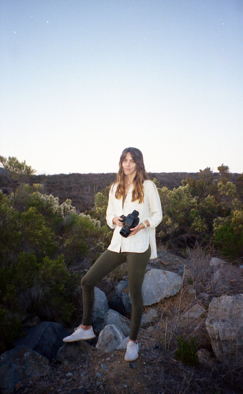 Destination photographer Standing in a natural setting with one leg propped up on some rocks, holding her medium format film camera. She is wearing a white collared shirt and has green pants on, and the image is taken on 35mm film