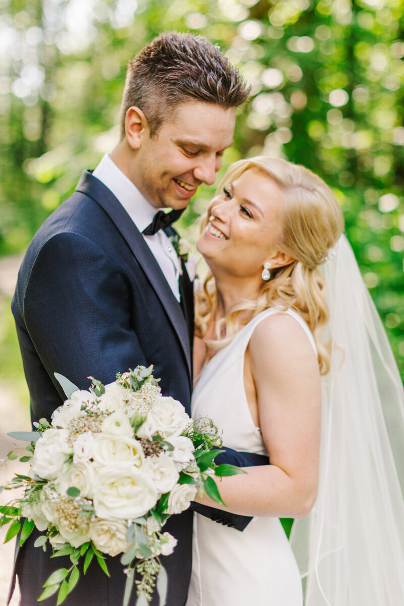 Bride and groom smiling with their arms around each other while bride holds white bouquet of flowers