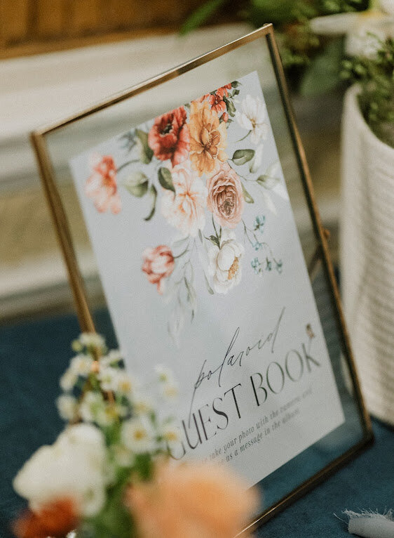 A dusty blue Guestbook Sign for a wedding welcome table features watercolour floral artwork in a gold frame is placed beside flowers at a wedding at Chateau Laurier in Ottawa Ontario.