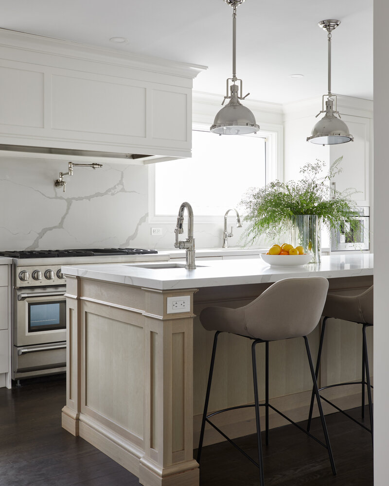 A modern luxury kitchen island lined with modern beige chairs in a bright kitchen area with white cabinetry and a white patterned backsplash