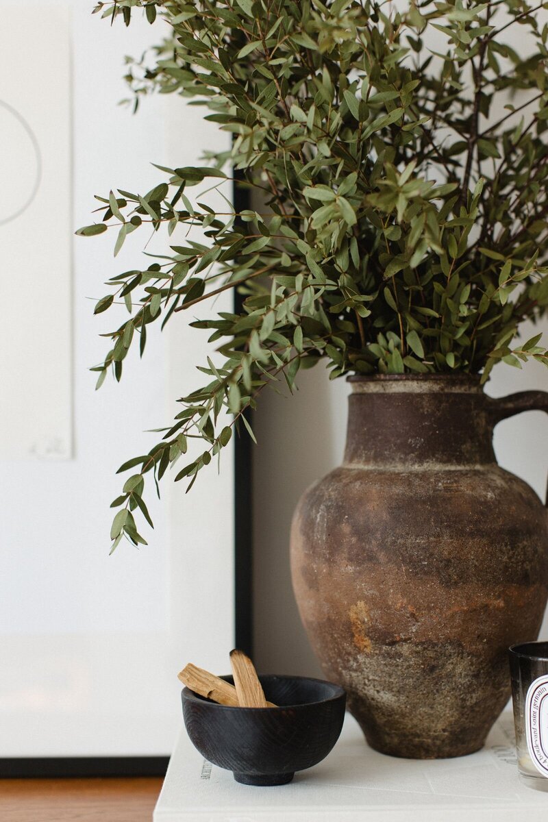 Close-up of a styled sideboard vignette featuring a brown antique vessel with greenery, a black wooden bowl with Santo Paolo and a Diptyque candle, all arranged on an Architectural Digest book, with a framed print in the background.