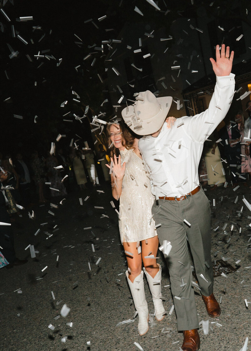 Flash photo of bride in white cowgirl boots and groom in cowboy hat leaving their reception surrounded by confetti