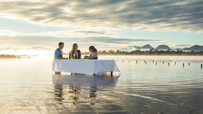 A group of diners enjoys freshly shucked oysters and local Tasmanian wine at a submerged dinner table in a tranquil coastal setting.