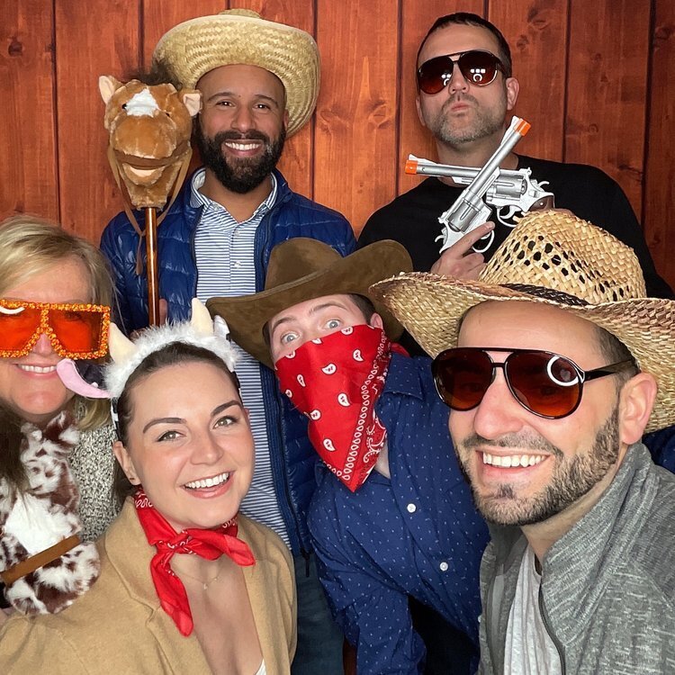 Executive team posing in front of a western wooden backdrop wearing western props like cowboy hats and bandanas — a customized western themed photo booth Phoenix Arizona solution for corporate events.