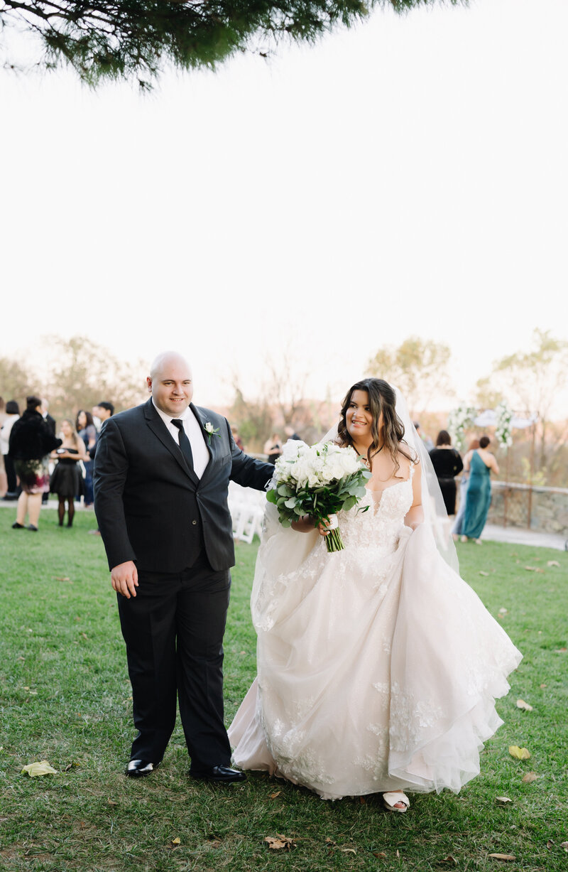 bride holding a white rose bouquet and carrying her wedding dress as her groom walks along side her and they smile at one another at Charlottesville wedding venues for an outdoor wedding