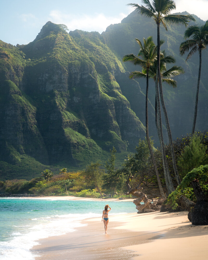 Woman in a blue swimsuit walking on a beach towards a green mountain