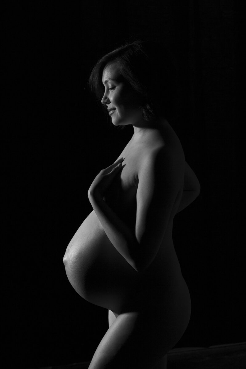 black and white nude portrait of an Asian mom to be on a black background private areas covered in shadow focus on the bare belly.  Charlotte Maternity Photographer NC created by Insley Photography