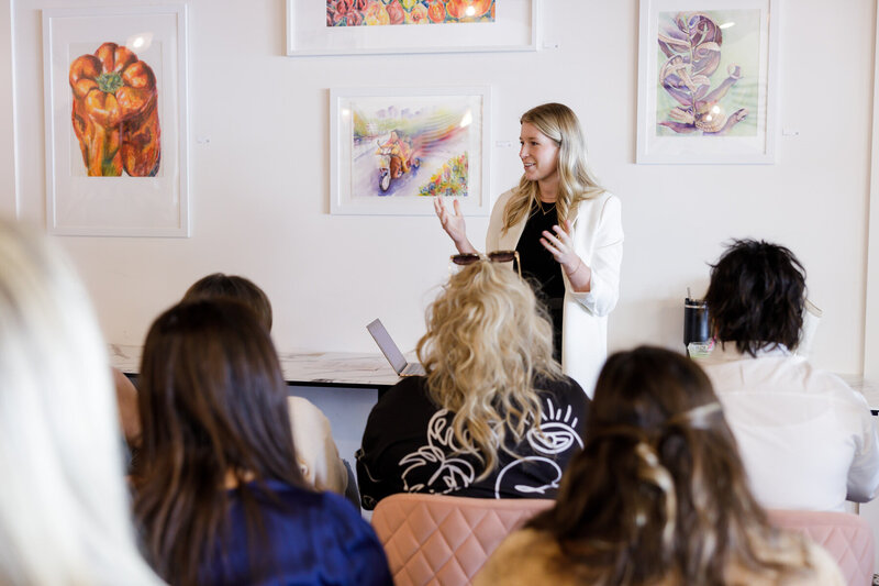 A woman stands in front of a small group, gesturing with her hands as she speaks. The audience's backs are to the camera. The room's walls are adorned with colorful artwork, and a small table is visible in the background. Everyone appears to be engaging in spirited networking, typical of Seattle's vibrant communities.