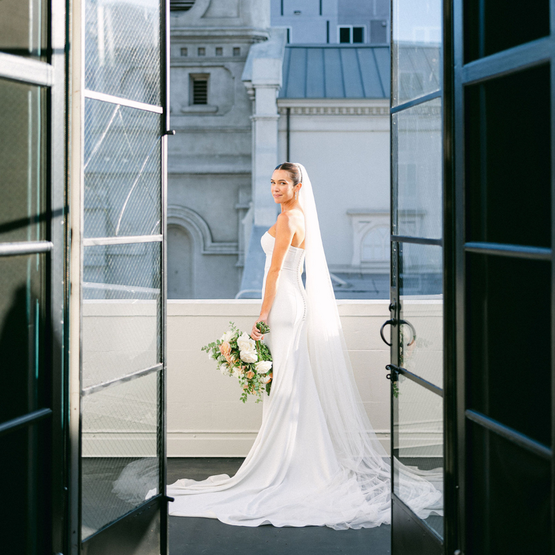 bride stands on the balcony at Vibiana and her portrait is taken through the doors overlooking the city of los angeles captured by los angeles wedding photographer magnolia west photography