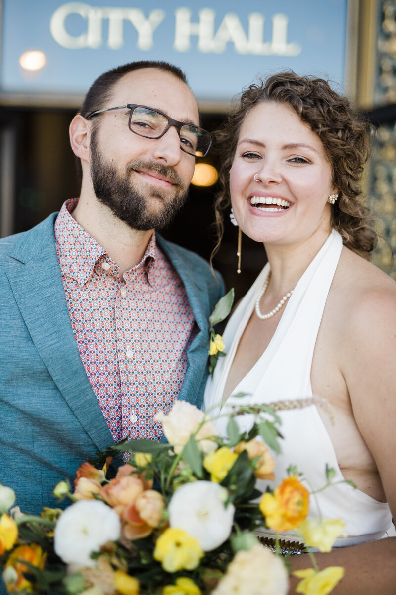 a bride and groom stanging right next to eachother, looking at the camera and laughing, a sign that reads "city hall" is visible behind them. The groom is wearing a very cool relazed suit jacket and button up shirt with no tie, the bride wears a halter jumpsuit