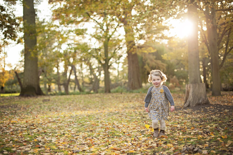 A young girl walks through the autumn leaves that have fallen on the ground and looks directly at the camera. Behind the low sun is shining through the golden leaves.