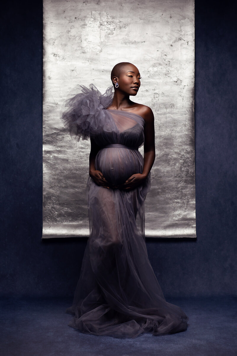 A striking maternity portrait of a dark-skinned pregnant woman wearing a sheer gray tulle gown with dramatic ruffled detailing on one shoulder. She poses confidently in front of a silver backdrop, captured by Dallas Maternity Photographer Lindsay Walden.